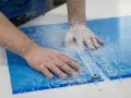 Hands guide a router, creating a clean cut on a blue plexiglass sheet, leaving a trail of white shavings. This is how you cut plexiglass with a router.
