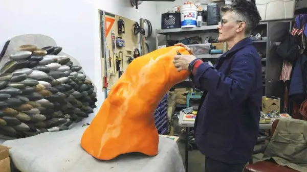 Artist applies finishing touches to a large, orange, 3D printed object, part of a project creating large objects.