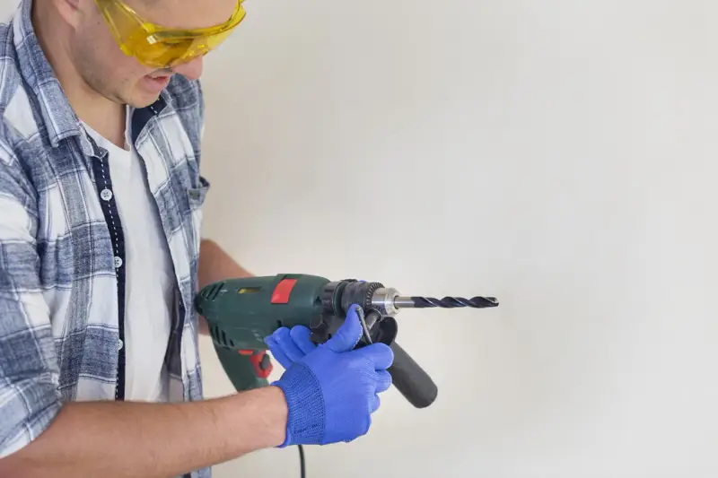 Man in safety glasses and blue gloves holding a drill to a white surface.