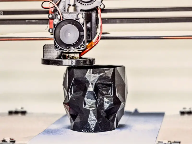A 3D printer nozzle extrudes filament to create a faceted black skull planter, showcasing innovative 3D printing examples.